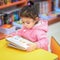 Little Girl Indoors In Front Of Books. Cute Young Toddler Sitting On A Chair Near Table and Reading Book. Library, Shop.