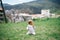 Little girl kneels on a green meadow with an apple in her hand against the background of houses