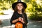 little girl with long dark hair smiles in a Halloween witch costume and holds a pumpkin-shaped candy bowl outside. Halloween for