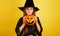 little girl with long hair smiles in a Halloween witch costume and holds a pumpkin-shaped candy bowl on a yellow background