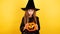 little girl with long hair smiles in a Halloween witch costume and holds a pumpkin-shaped candy bowl on a yellow background
