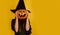 little girl with long hair smiles in a Halloween witch costume and holds a pumpkin-shaped candy bowl on a yellow background. Copy