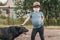 Little girl in a medical mask stroking a large homeless black dog outdoors