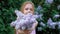 A little girl outdoors in a park or garden holds lilac flowers. Lilac bushes in the background. Summer, park