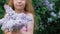 A little girl outdoors in a park or garden holds lilac flowers. Lilac bushes in the background. Summer, park