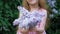 A little girl outdoors in a park or garden holds lilac flowers. Lilac bushes in the background. Summer, park