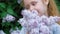 A little girl outdoors in a park or garden holds lilac flowers. Lilac bushes in the background. Summer, park