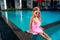 Little girl in pink dress and funny sunglasses sits by poolside. Child enjoys sunny day at outdoor swimming pool, summer