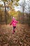 Little girl in pink jacket, knit cap and resin boots happily running in autumn forest on golden leaves cover backwards to camera.