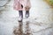 Little girl in pink waterproof raincoat, purple rubber boots funny jumps through puddles on street road in rainy day