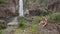 Little Girl Plays on Stone against High Waterfall Cascade