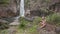 Little Girl Plays on Stone against High Waterfall Cascade