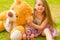 Little girl preschool sitting on backyard next to her teddy bear, wearing her roller skates and crossing her legs, in a