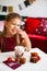 Little girl in red dress eating Christmas cookies with cacao in cup, red Chirstmas decorations around