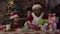 A little girl in a red and white suit sits next to Santa Claus and gives him a letter in an envelope