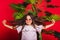 A little girl with round sunglasses stands next to a palm tree on a solid red background. A cheerful and energetic child