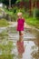 Little girl in rubber boots stands in the middle of a dirty large deep puddle on a country sandy road after rain