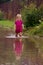 Little girl in rubber boots stands in the middle of a dirty large deep puddle on a country sandy road after rain