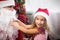 Little girl in Santa hat on Santa`s lap near Christmas tree in Christmas decor. Shows a box with a gift, a soft toy, touches his b