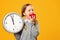 Little girl schoolgirl holds a big clock and bites a red apple on a yellow background. Break and lunch