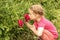 A little girl sits by a flower bed in the garden and sniffs bright pink peonies