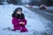 A little girl sits in the snow by the road and watches passing cars