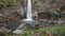 Little Girl Sits on Stone against High Waterfall Cascade