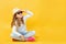 Little girl sitting on the floor in the studio with a hat and sunglasses on a yellow background.