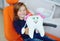 a little girl is sitting smiling in an orange dental chair and holding a large toy tooth in her hands.