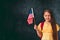 Little girl smiling and holding American flag standing  standing against blackboard , copy space, english language studying