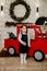 A little girl stands near a red toy car against the background of a Christmas wreath made of spruce
