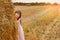 A little girl in a sundress stands in a field behind a haystack of mown grass, on a sunny day. Children in nature. Lifestyle