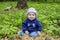 A little girl with sweater and hat sits in the middle of the forest and looks into the camera. A happy girl learns the world,