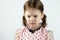 A little girl with two ponytails in a dress with polka dots on a white background looks down angrily. Studio photo