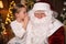 Little girl whispering in Santa Claus` ear near Christmas tree indoors
