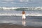Little Girl in White Dress Standing at Ocean Edge as Wave Rolls In