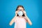 Little girl wiping foggy glasses caused by wearing disposable mask on blue background. Protective measure during coronavirus