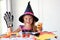 A little girl in a witch costume looks at the camera and laughs, holding a glass pumpkin and a skeleton glove in her hands