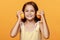 A little girl in a yellow dress, on an orange background, makes headphones out of orange halves.
