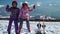 Little girls in outwear and masks with helmet sitting on wood fence with skis near against mountains