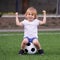 Little goalkeeper: blonde toddler kid sitting on soccer ball at green football stadium jokingly showing bicepses. Success concept