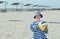 A little happy boy standing on the beach holds a bright soccer ball against the background of umbrellas from the sun and the sea,