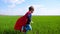 A little happy child in a superhero costume, a red cloak and a mask runs on green grass against a blue sky