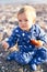 Little kid in a blue overalls sits on a pebble beach, clutching an apple in his hand. Close-up