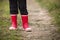 Little kid boy in rain rubber boots. leg`s shot. Child in bright red shoes. On a countryside road, outdoors