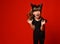 Little kid with devil horns, in black blouse and leggings screaming with closed eyes, posing against red background. Close up