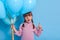 Little kid with pigtails bunch of balloon and pointing aside with fore finger, keeps mouth opened, looks aside with interest,