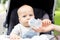 Little kid sitting in stroller and drinking water from bottle during walk at hot summer day. Mom feeding infant boy with drink. Da