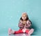 Little lady in headband, pink faux fur coat, pants and boots. She smiling, sitting on floor against blue background. Close up