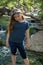 Little latina girl smiling coyly and looking down while standing in front of a stream and woods in the shade